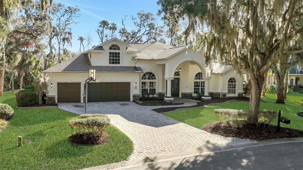 A beige house with a paved driveway, a three-car garage, and large oak trees — 11 CYPRESS HOLLOW LANE, ORMOND BEACH, FL