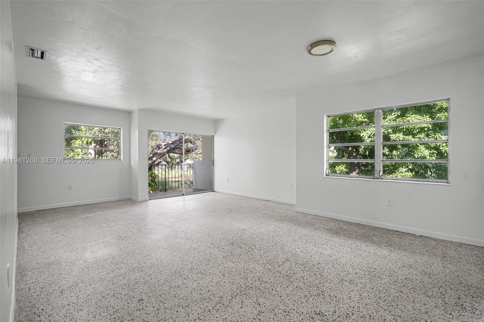 An empty room features white walls, a speckled floor, and windows overlooking green trees — 11330 Peachtree Dr, Miami FL 33161, MIAMI, FL