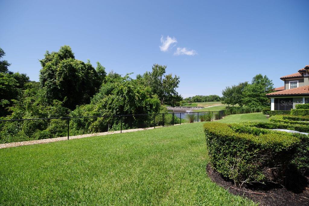 A grassy backyard features a black metal fence, lush landscaping, and a view of water — 1541 CORKERY COURT UNIT 1541, WINTER SPRINGS, FL