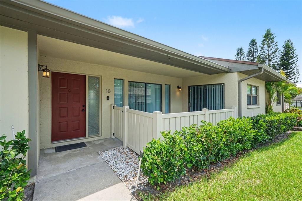 A red front door with a white fence and landscaping sits under a covered porch — 10 LESLEY LANE, OLDSMAR, FL