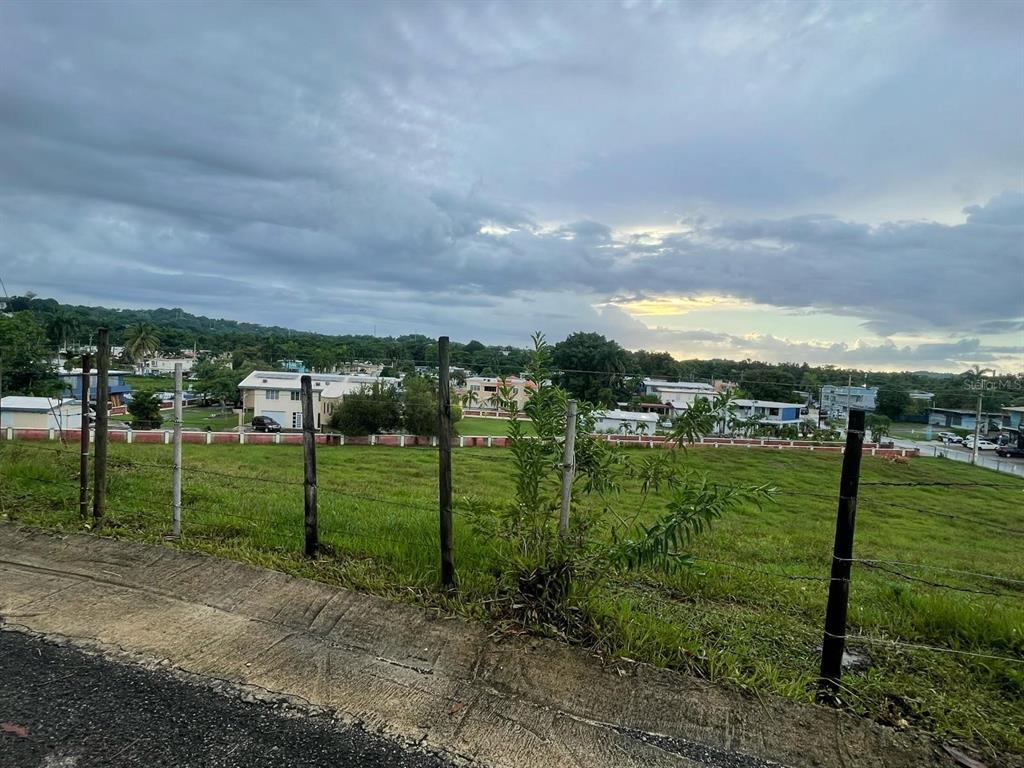 A grassy, fenced-in vacant lot overlooks a residential neighborhood under a cloudy sky — PR-417 BARRIO MARIAS EN AGUADA, AGUADA, PR