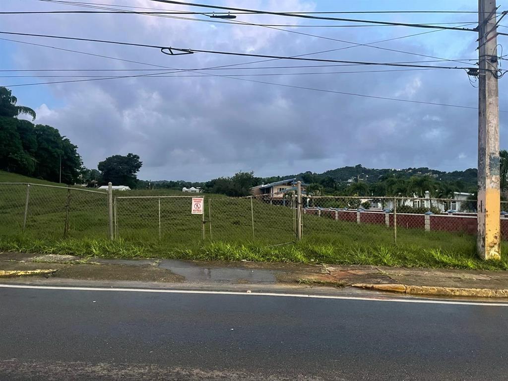 A fenced, grassy vacant lot sits along a paved road under a cloudy sky — PR-417 BARRIO MARIAS EN AGUADA, AGUADA, PR