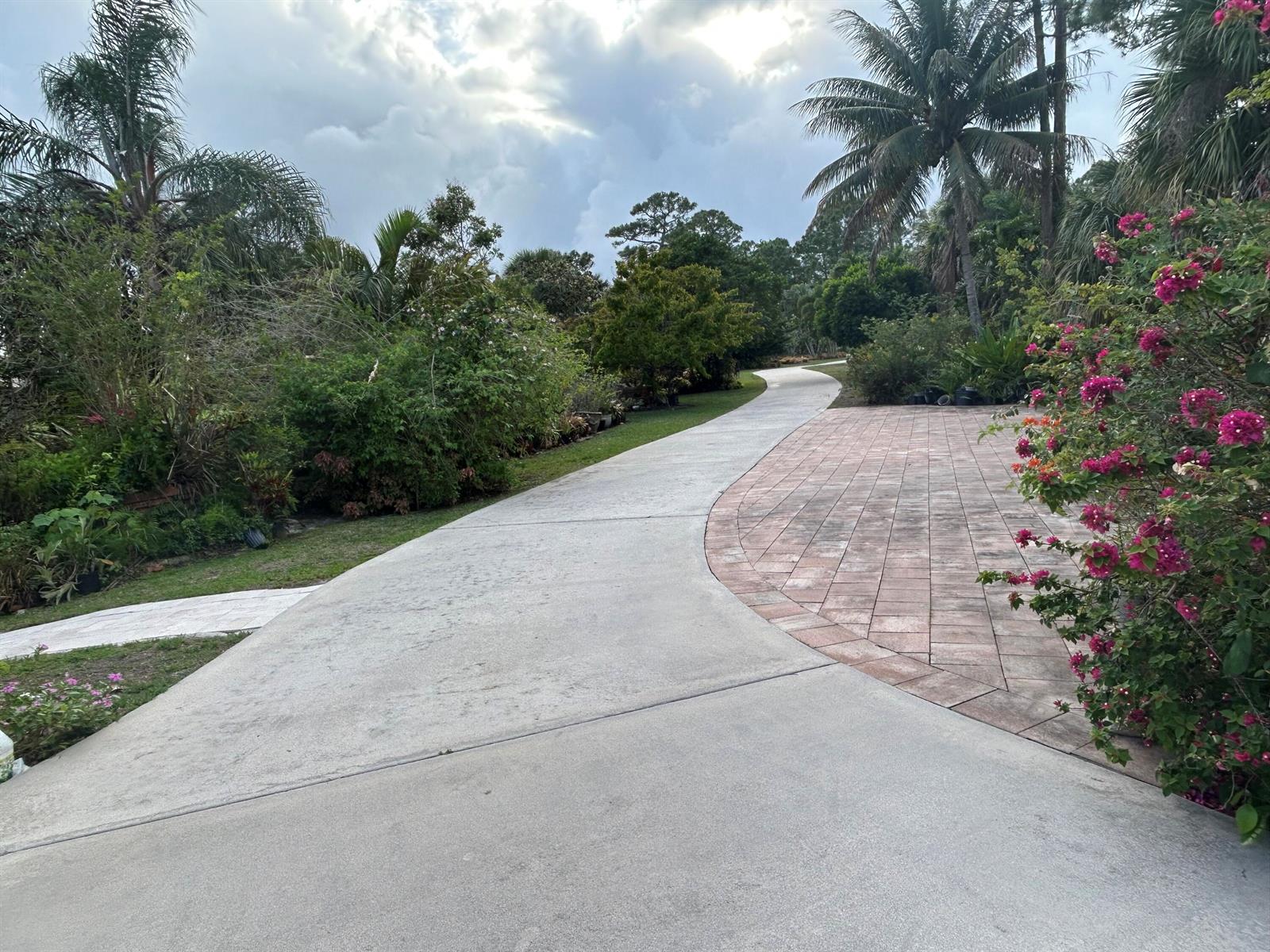 A concrete driveway curves past a brick-paved area and lush landscaping under a cloudy sky — 6220 Kelty Way, Lake Worth FL 33467, LAKE WORTH, FL