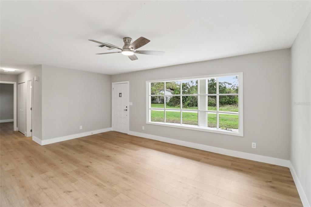 A bright living room features light wood-look flooring, gray walls, and a large grid window — 1412 DIGARMO TERRACE, DELTONA, FL