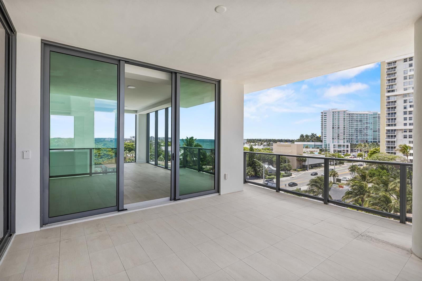 An outdoor balcony with light-colored tile flooring, glass railings, and sliding doors overlooking a city — 305 Briny Avenue # 506, Pompano Beach FL 33062, POMPANO BEACH, FL