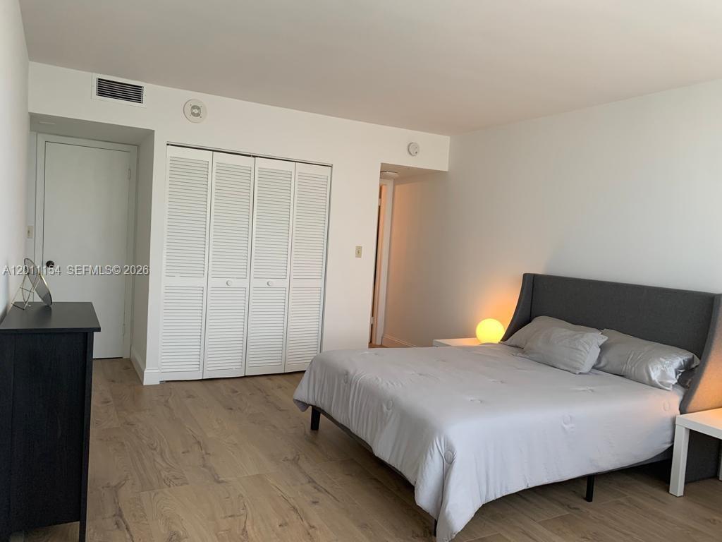 A bedroom features light wood-look flooring, a gray upholstered bed, and white louvered closet doors — 780 NE 69th St # 506, Miami FL 33138, MIAMI, FL