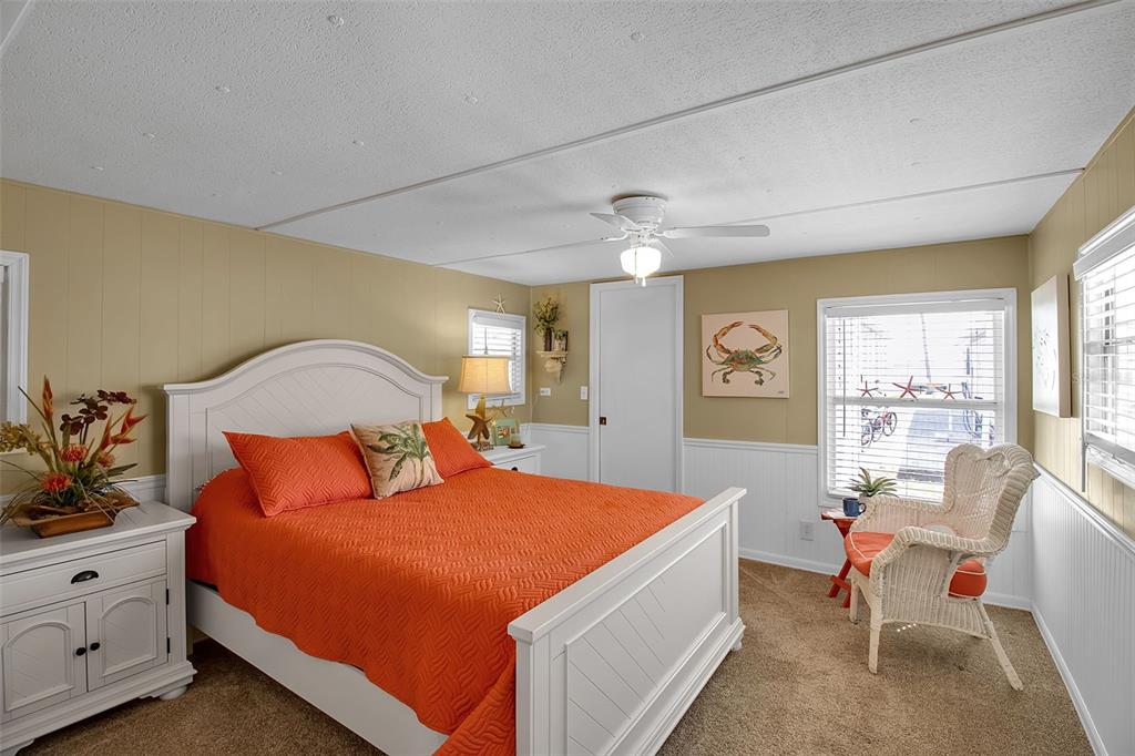 A bedroom features a white bed with an orange quilt, beige walls, and a ceiling fan — 10315 CORTEZ ROAD W UNIT 27F, BRADENTON, FL
