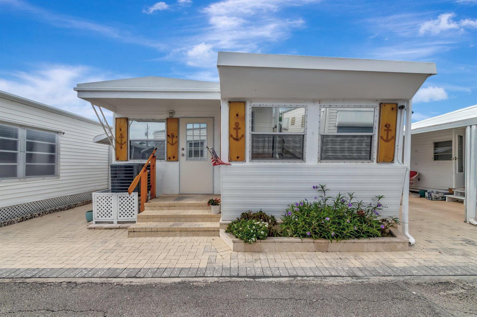 A white mobile home with anchor-themed shutters, a paved patio, and a small garden bed — 18 Juniper Drive # J, Briny Breezes FL 33435, BRINY BREEZES, FL