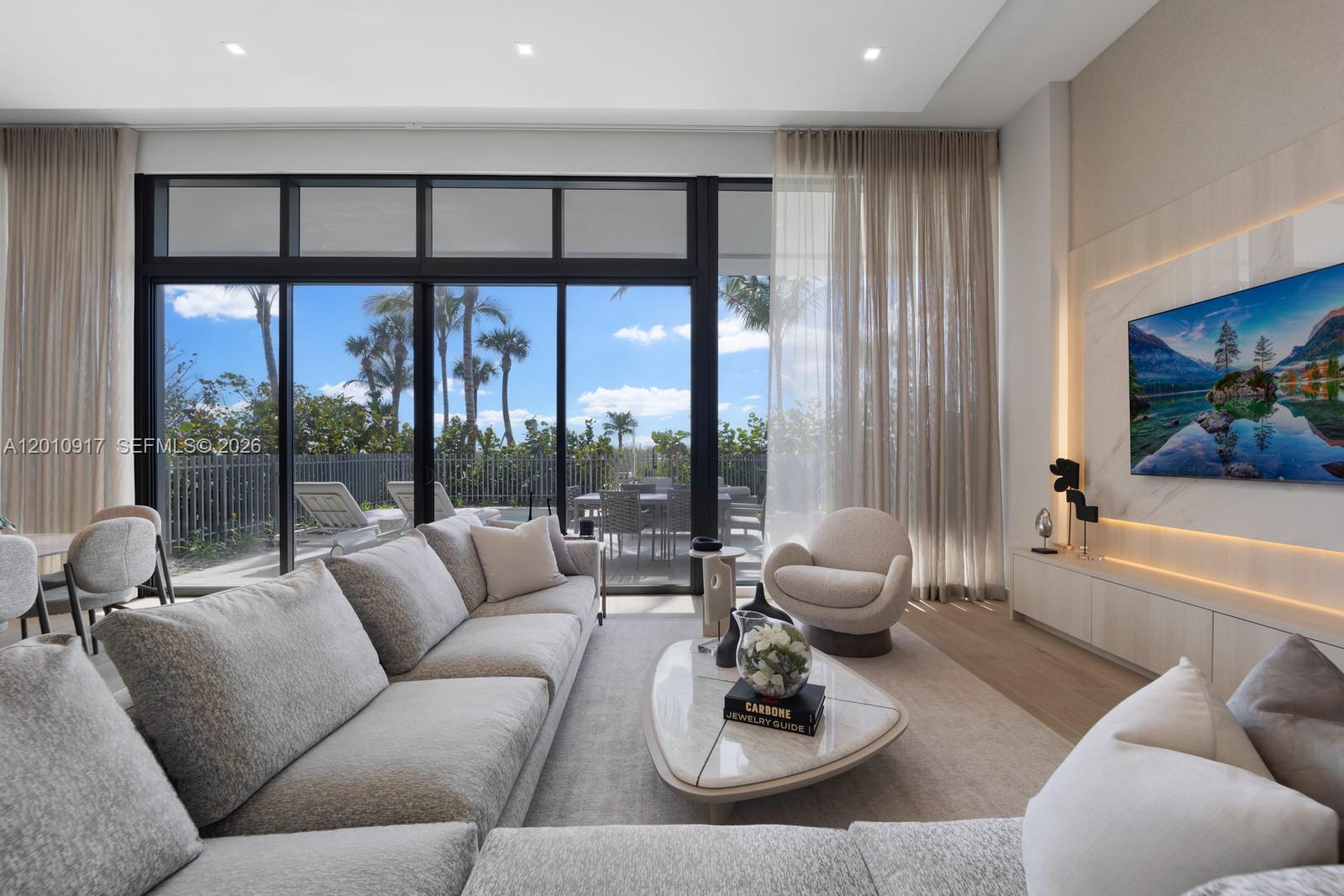 A modern living room features a wall-mounted television, a textured accent wall, and neutral furniture — 900 N Ocean Blvd # 106, Pompano Beach FL 33062, POMPANO BEACH, FL