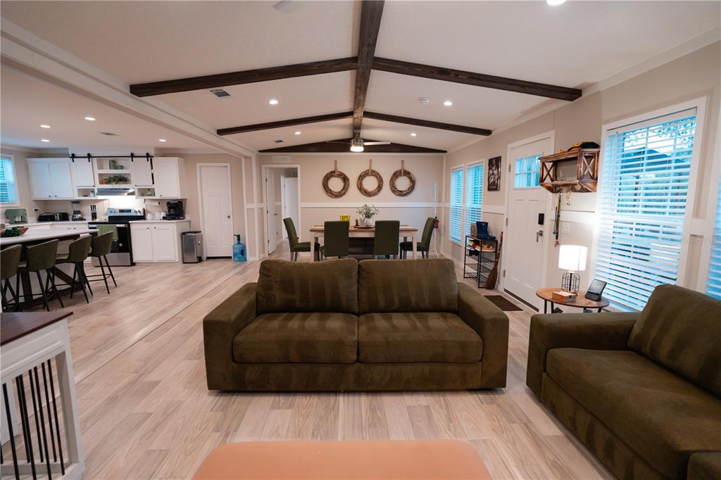 An open-concept living room and kitchen feature wood-look flooring, exposed ceiling beams, and neutral walls — 7670 NE 97TH COURT, BRONSON, FL
