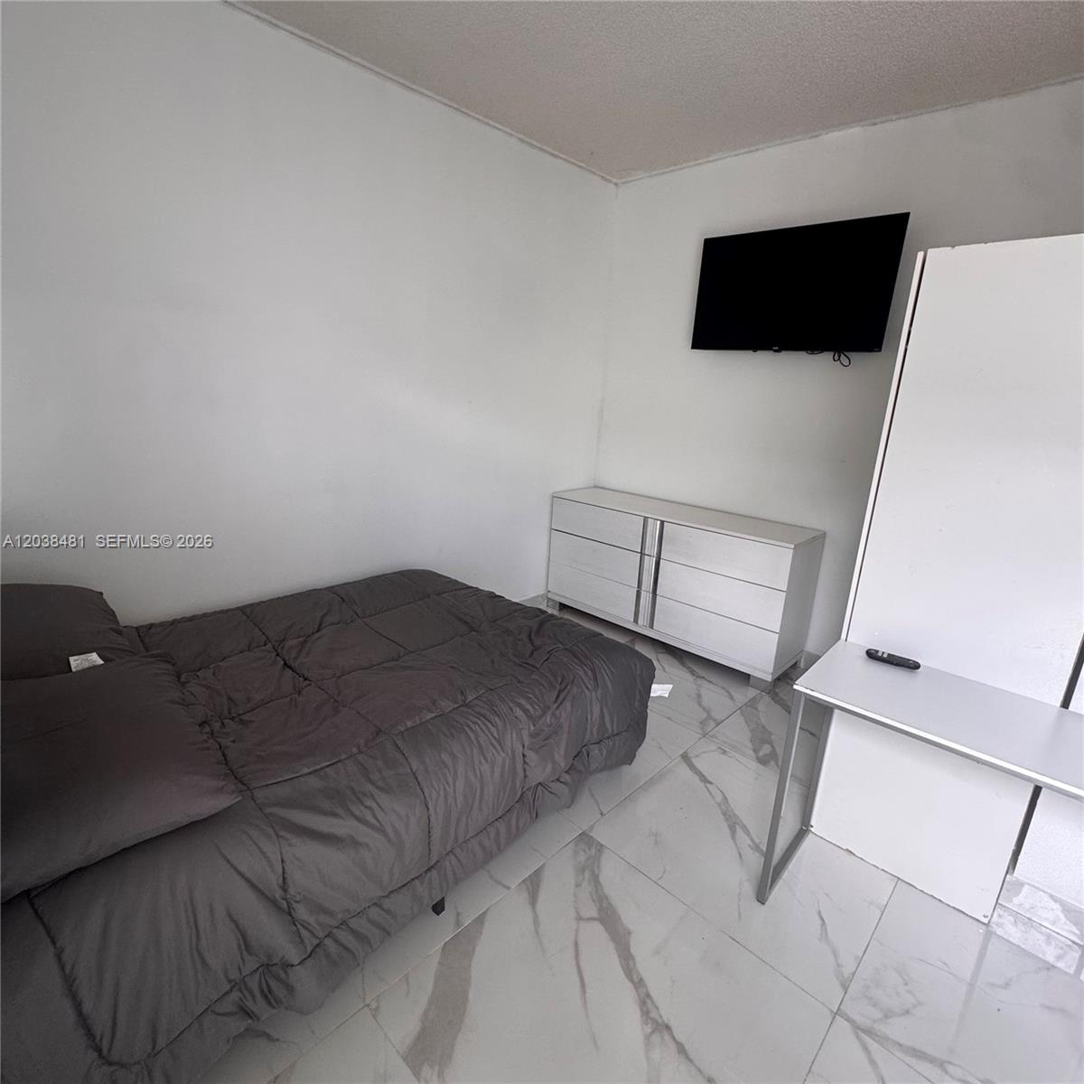 A bedroom features a bed, a white dresser, a wall-mounted television, and marble-patterned tile flooring — MIAMI, FL