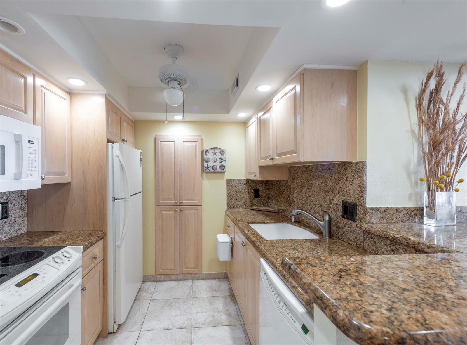 A galley kitchen features light wood cabinets, granite countertops, white appliances, and tile flooring — 111 Briny Avenue # 12, Pompano Beach FL 33062, POMPANO BEACH, FL