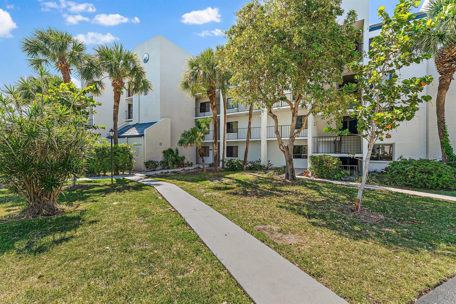 A concrete walkway leads toward a multi-story apartment building surrounded by palm trees and landscaping — 1605 S Us Highway 1 # B406, Jupiter FL 33477, JUPITER, FL
