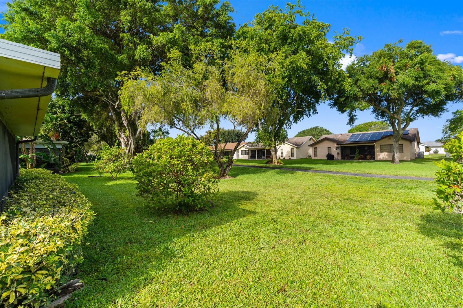 A grassy backyard with mature trees, shrubs, and neighboring houses under a clear blue sky — 18051 S 104th Ter Terrace S, Boca Raton FL 33498, BOCA RATON, FL