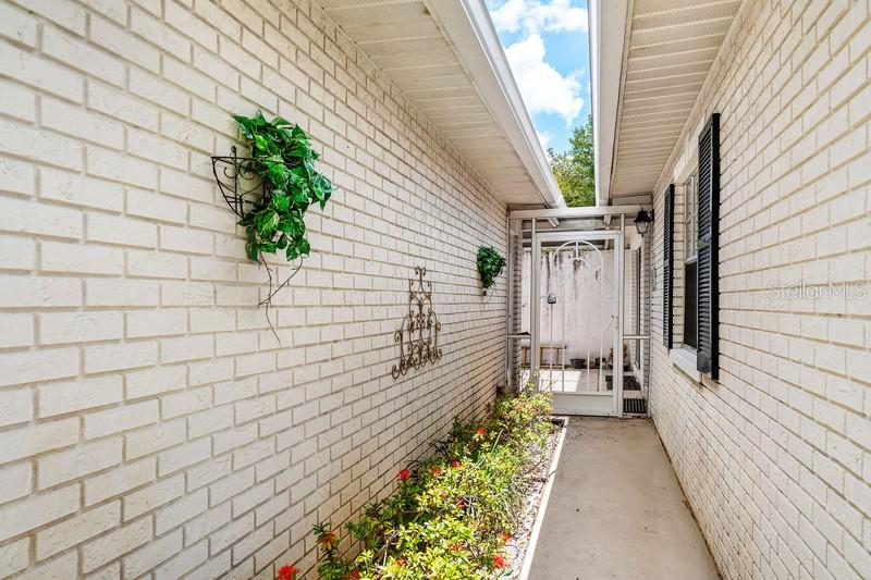 A narrow outdoor walkway with white brick walls, a small garden bed, and a screen door — 10116 S 44TH AVENUE S UNIT 328, BOYNTON BEACH, FL