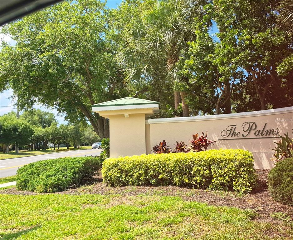 A beige stucco monument sign for The Palms stands near a road and trees — 4802 51ST STREET W UNIT 1116, BRADENTON, FL