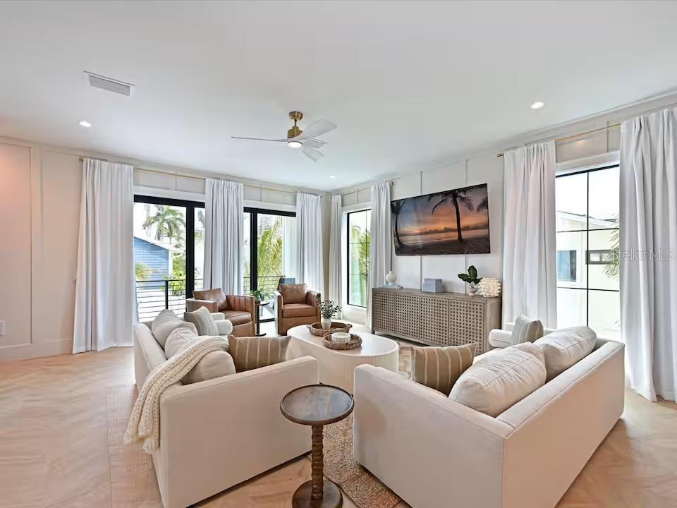 A bright living room features light wood floors, white walls, two sofas, and large windows — 2209 AVENUE B, BRADENTON BEACH, FL