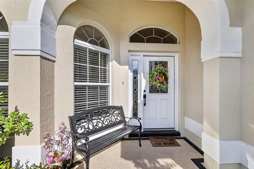 A covered entryway features a white front door, a black metal bench, and a doormat — 17325 SE 73RD PARK GLEN COURT, THE VILLAGES, FL