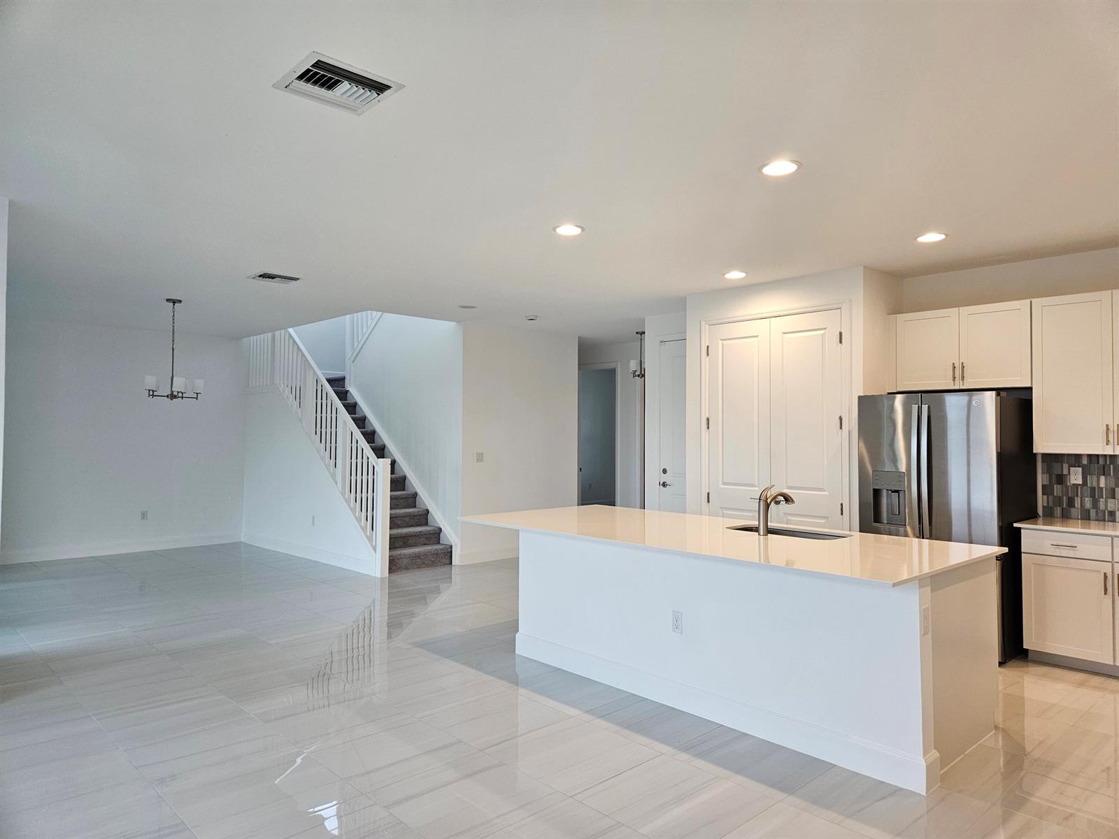 A modern kitchen with a white island and stainless steel refrigerator overlooks a staircase area — 17199 Tawny Owl Trail, FL 33470, FL