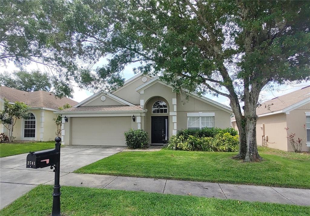 A single-story suburban house with a beige stucco exterior, a two-car garage, and a tree — 2741 BELLEWATER PLACE, OVIEDO, FL