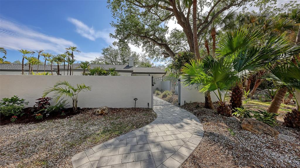 A paved walkway leads to a metal gate in a white wall surrounded by landscaping — 7345 PINE NEEDLE ROAD, SARASOTA, FL