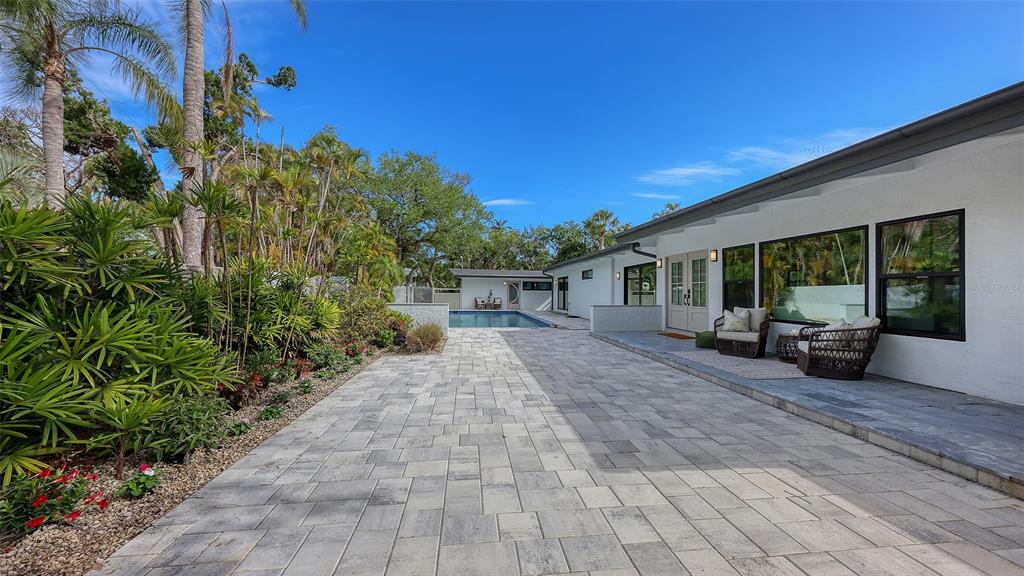 A paved patio area leads to a swimming pool beside a white house with large windows — 7345 PINE NEEDLE ROAD, SARASOTA, FL
