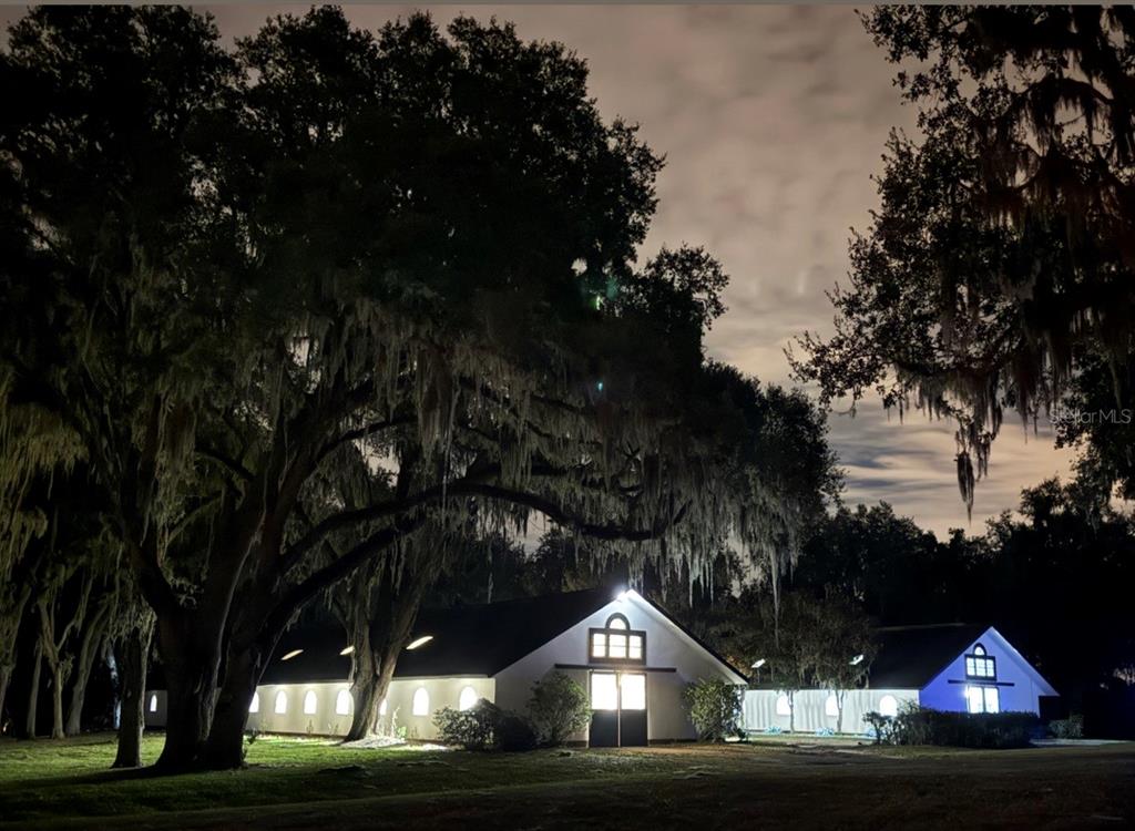 Two illuminated white buildings with arched windows sit under large oak trees at night — 5540 SE 157TH STREET ROAD, SUMMERFIELD, FL