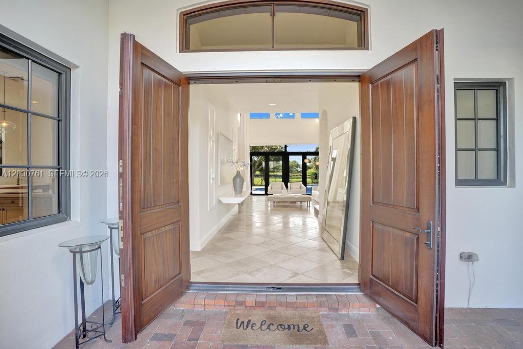 A view through open wooden double doors into a bright, tiled entryway with high ceilings — 10292 Emerson St, Parkland FL 33076, PARKLAND, FL
