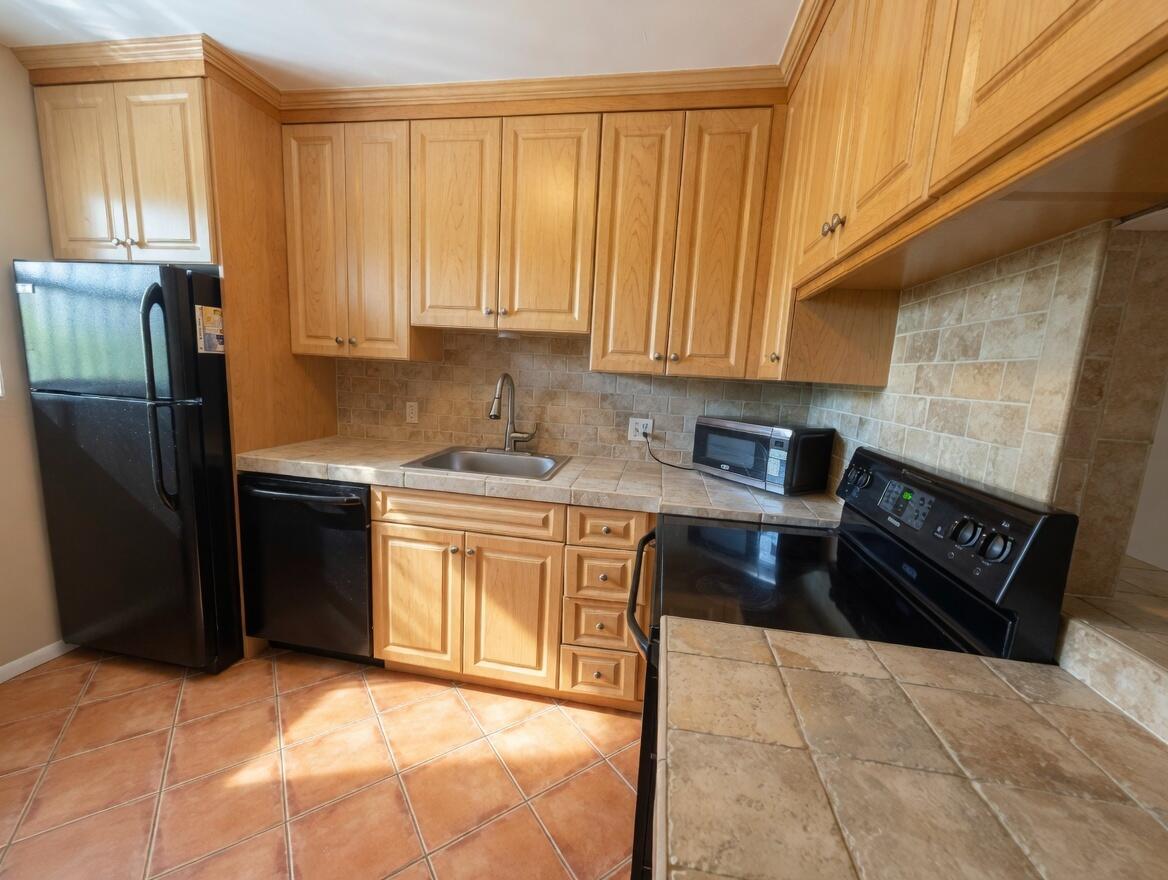 A kitchen featuring light wood cabinets, black appliances, and tan tile countertops and flooring — 1430 S Lakeside Drive # 22, FL 33460, FL