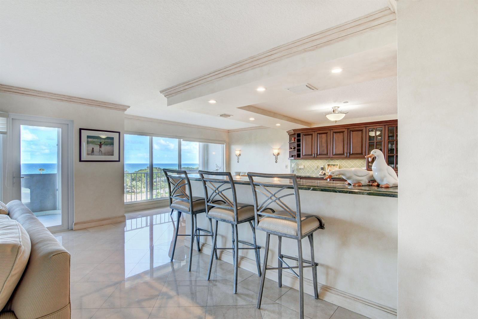 A kitchen bar area features three metal stools, dark wood cabinets, and ocean views — 4301 N Ocean Boulevard # 607, Boca Raton FL 33431, BOCA RATON, FL