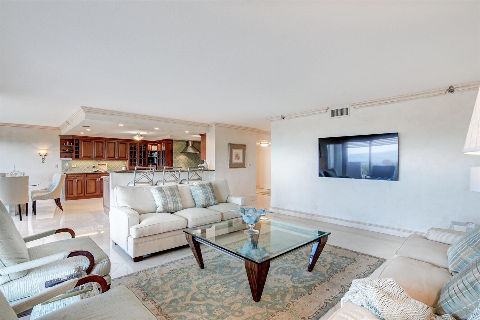 A living room with a glass coffee table, beige sofas, and an open-concept kitchen — 4301 N Ocean Boulevard # 607, Boca Raton FL 33431, BOCA RATON, FL