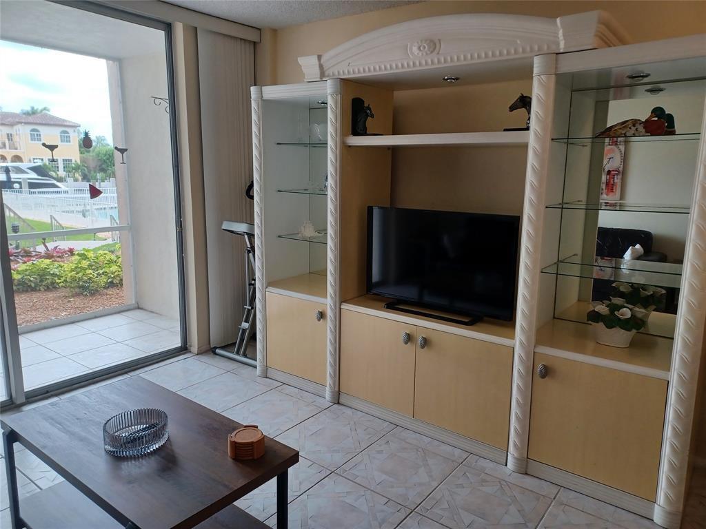 A living room features a large entertainment center, a coffee table, and a sliding glass door — 1391 S Ocean Boulevard # 105, Pompano Beach FL 33062, POMPANO BEACH, FL