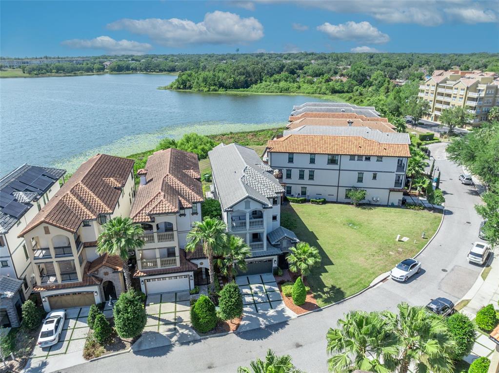 An aerial view shows several multi-story townhomes situated along a lake with a paved street — 7613 TOSCANA BOULEVARD, ORLANDO, FL