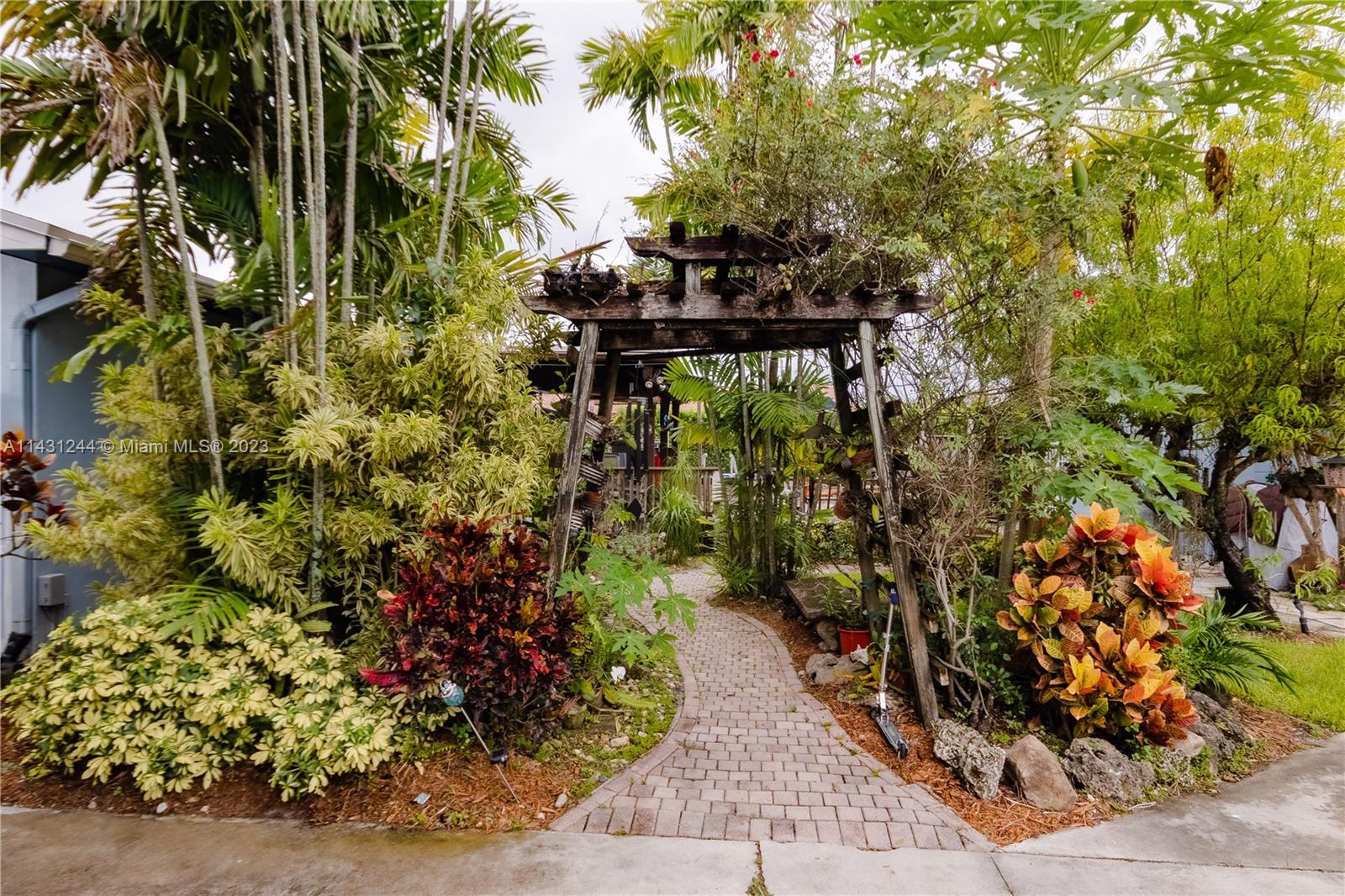 A brick pathway leads through a wooden arbor surrounded by lush tropical plants and trees — 2533 Fillmore St, Hollywood FL 33020, HOLLYWOOD, FL