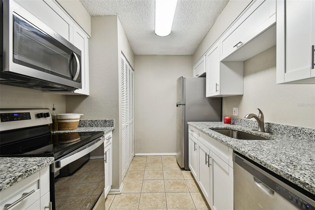 A galley kitchen features white cabinets, granite countertops, stainless steel appliances, and beige tile flooring — 7504 PRESLEY PLACE UNIT 115 C, TAMPA, FL
