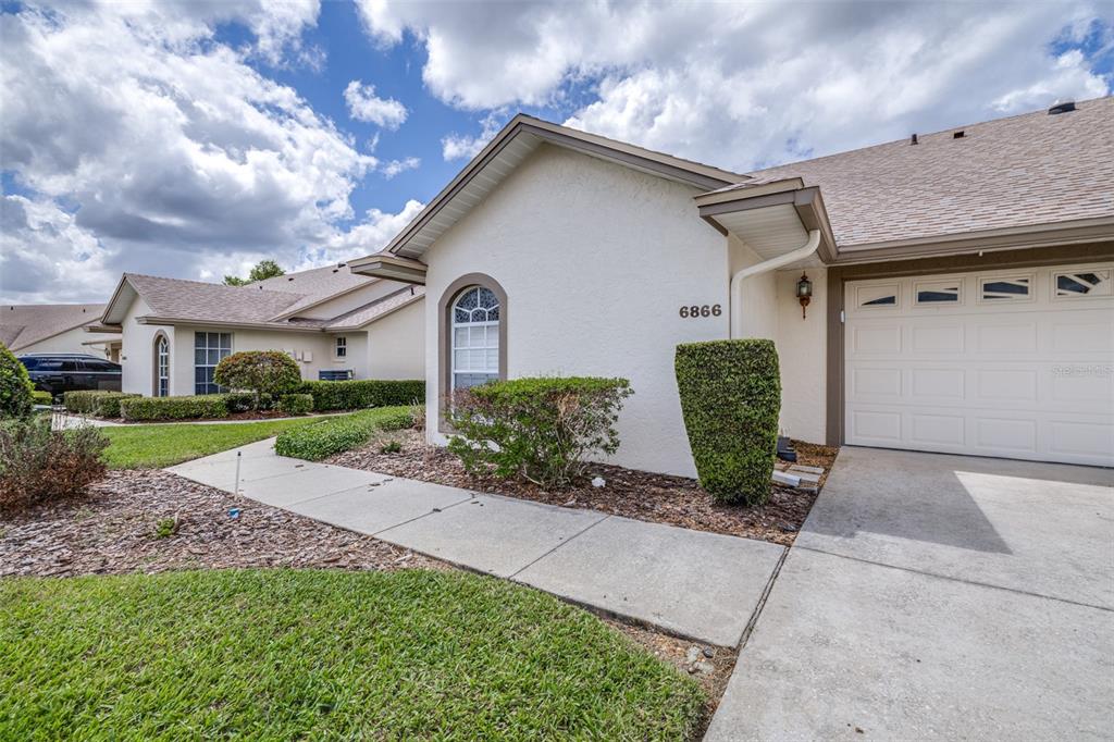 A single-story house exterior features a beige stucco facade, a garage, and a concrete walkway — 6866 BENDELOW DRIVE, LAKELAND, FL