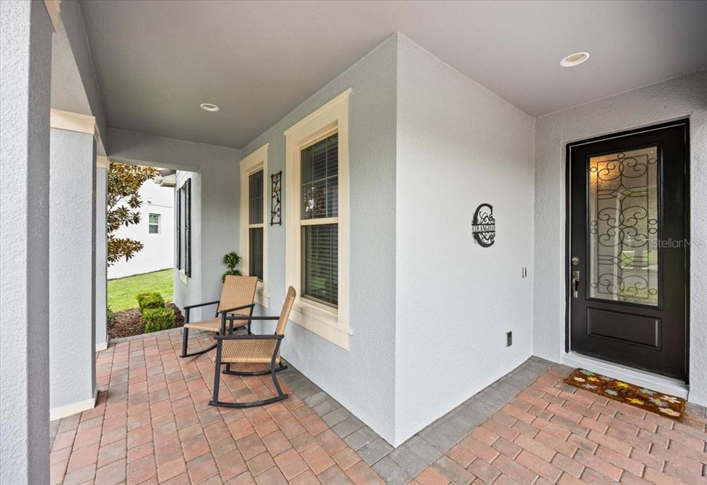 A covered front porch with brick pavers, two rocking chairs, and a black front door — 10043 LITTLE ALCOVE LOOP, WINTER GARDEN, FL