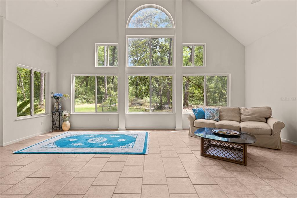 A spacious living room with vaulted ceilings, tile flooring, and large windows overlooking wooded scenery — 8250 WAUKEENAH HIGHWAY, MONTICELLO, FL