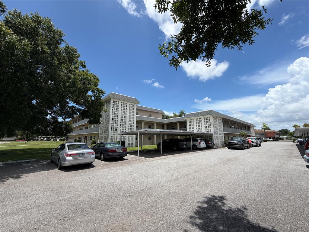 A multi-story apartment building features covered parking spaces in a paved lot under a blue sky — 5725 12TH AVENUE N UNIT 306D, ST PETERSBURG, FL