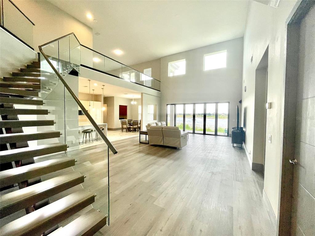A modern living room features a floating staircase, glass railings, and large windows overlooking water — 7759 GREEN MOUNTAIN WAY, WINTER GARDEN, FL