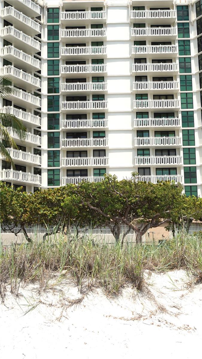 A tall white residential building with balconies stands behind sand dunes and green trees — 1111 Crandon Blvd # B705, Key Biscayne FL 33149, KEY BISCAYNE, FL