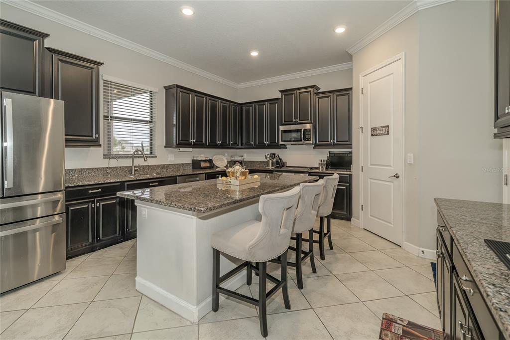 A kitchen with dark cabinets, granite countertops, a central island with seating, and stainless appliances — 8099 LAGO MIST WAY, WESLEY CHAPEL, FL