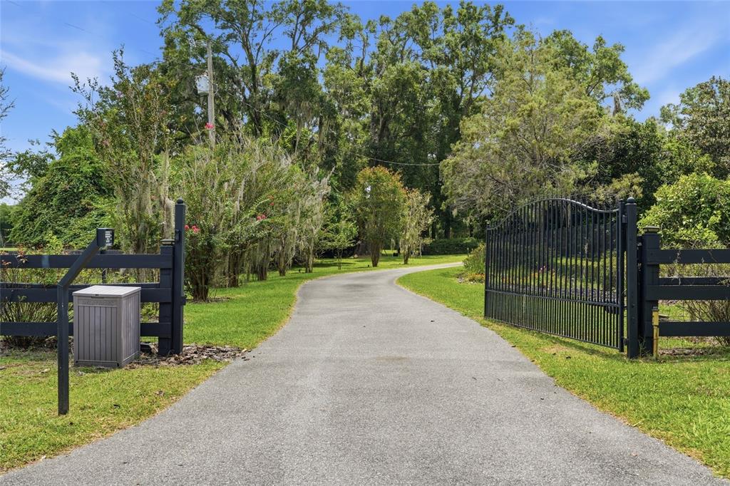 A paved driveway leads through an open black metal gate into a lush, tree-lined property — 26341 NW 166TH AVENUE, HIGH SPRINGS, FL