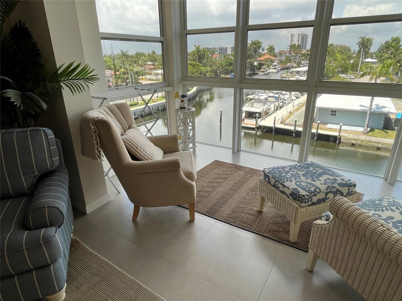 A sunroom features a beige armchair, patterned ottomans, and floor-to-ceiling windows overlooking a boat canal — 1481 S Ocean Boulevard # A, Pompano Beach FL 33062, POMPANO BEACH, FL