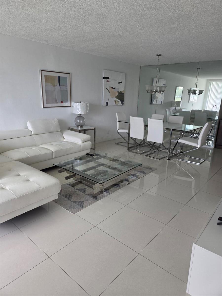 A modern living and dining area features white tile floors, a sectional sofa, and mirrors — 3091 N Course Drive # 208, Pompano Beach FL 33069, POMPANO BEACH, FL