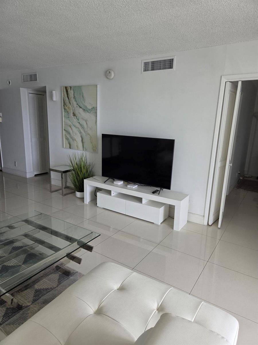 A living room with a white media console, flat-screen television, and white tile flooring — 3091 N Course Drive # 208, Pompano Beach FL 33069, POMPANO BEACH, FL