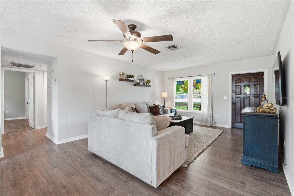 A living room features light gray walls, wood-look flooring, a ceiling fan, and a sofa — 2140 NE COACHMAN ROAD, CLEARWATER, FL