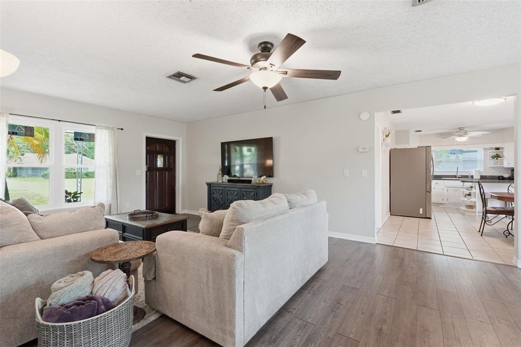 A living room with light gray walls, wood-look flooring, a ceiling fan, and an adjacent kitchen — 2140 NE COACHMAN ROAD, CLEARWATER, FL