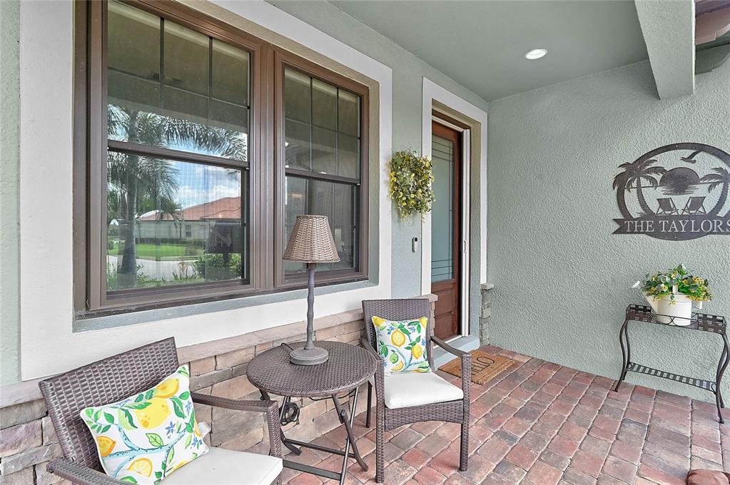 A covered porch features brick flooring, two wicker chairs with lemon pillows, and a small table — 20275 GRANLAGO DRIVE, VENICE, FL