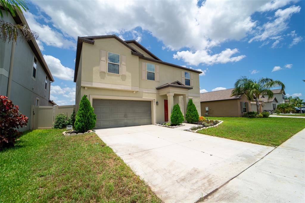 A two-story beige house features a two-car garage, a red front door, and landscaping — 9831 LEMON DROP LOOP, RUSKIN, FL